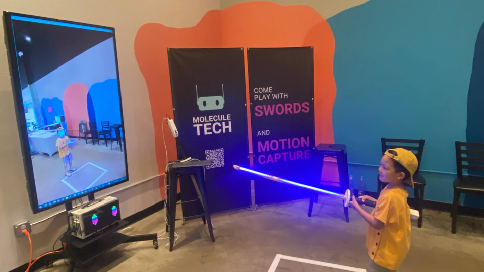 A young visitor holding an illuminated bokken beside the display and Molecule banner.